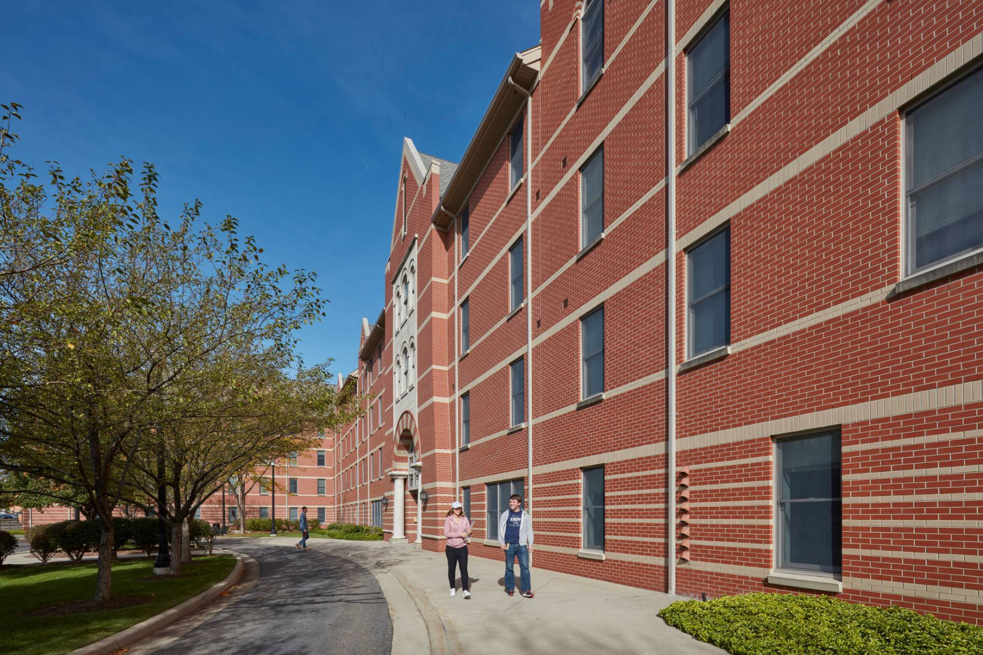 Winter Hall Living Center is a red-brick building with several windows on a sunny day. Two people walk along the sidewalk, with lush trees lining the path, creating a peaceful atmosphere.